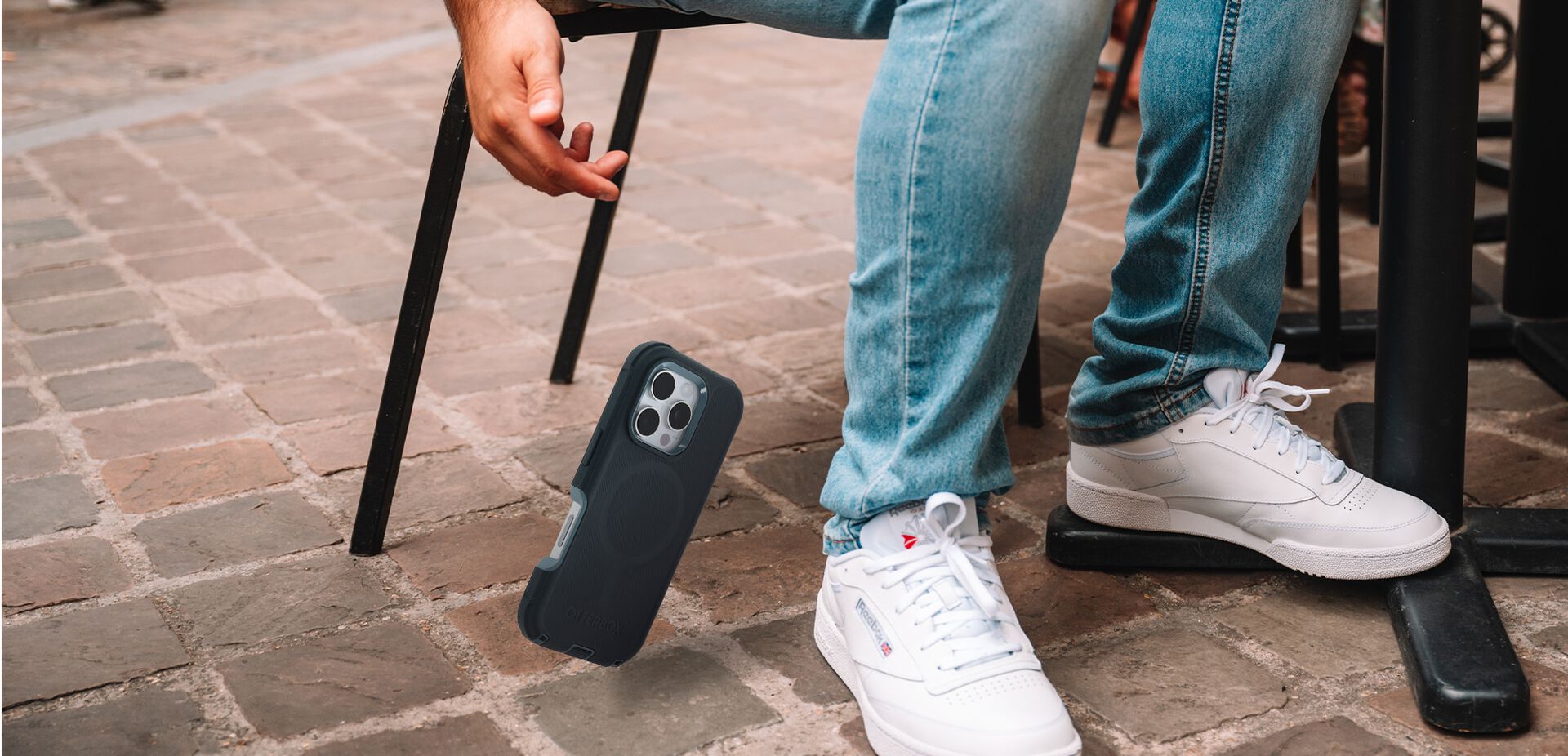 Person wearing white sneakers and blue jeans sitting on a chair with a phone case on the ground.